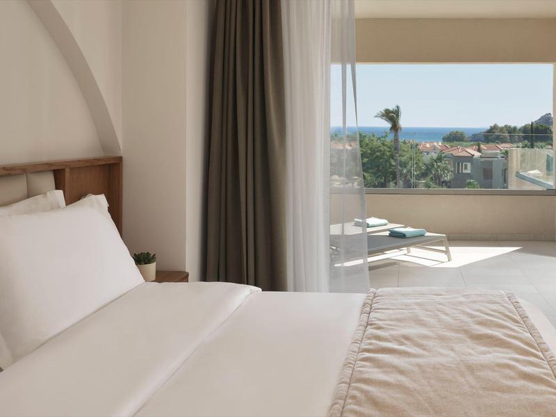 Camera d'hotel luminosa con letto bianco, tenda grigia e terrazza con vista mare.