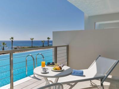 Balcone con piscina, vista mare, tavolo, bevande e sedia a sdraio sotto il cielo azzurro.
