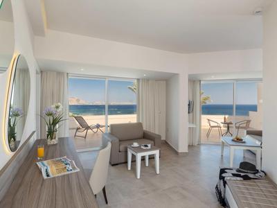 Suite d'hotel luminosa con zona soggiorno, scrivania e balcone con vista mare.