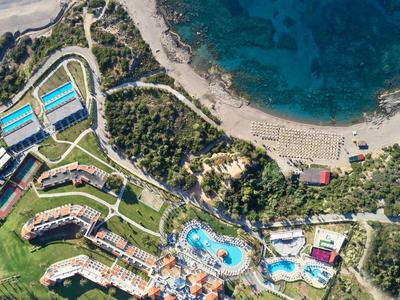 Veduta aerea di una località balneare con piscine, edifici, aree verdi e una spiaggia vicino a un'acqua azzurra cristallina.