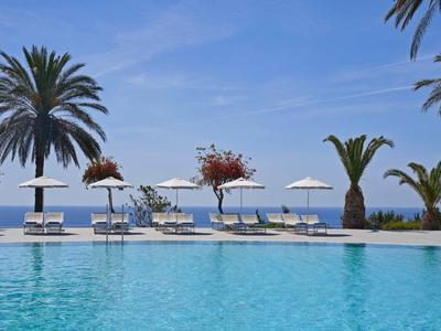 Pool mit Liegestühlen und Sonnenschirmen neben Palmen am Meer unter blauem Himmel.