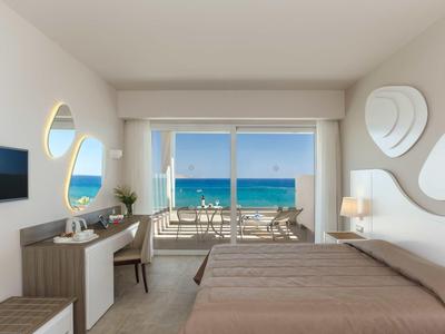 Camera d'hotel con letto, scrivania, TV e balcone con vista sull'oceano in una giornata di sole.