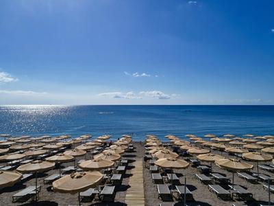 Strand mit Reihen von Sonnenschirmen und Liegen vor ruhigem blauem Meer unter sonnigem Himmel.