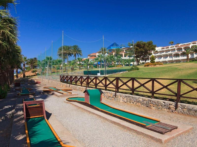 Campo da minigolf all'aperto con prato verde, recinzioni in legno ed edifici sullo sfondo.