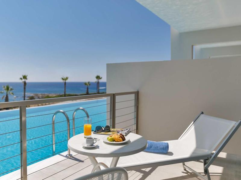 Balcone con piscina, vista mare, tavolo, bevande e sedia a sdraio sotto il cielo azzurro.