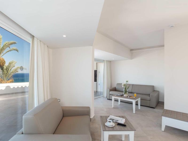 Lounge d'hotel moderna con divani beige, tavoli bianchi e ampia finestra con vista su palme.