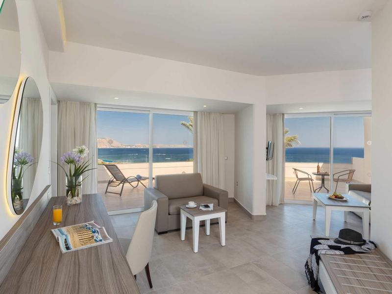 Suite d'hotel luminosa con zona soggiorno, scrivania e balcone con vista mare.