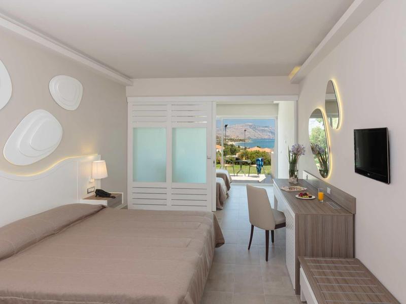 Camera d'hotel moderna con due letti, scrivania, sedia, TV a muro e balcone con vista mare.