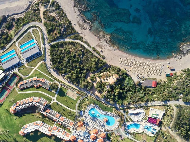 Veduta aerea di una località balneare con piscine, edifici, aree verdi e una spiaggia vicino a un'acqua azzurra cristallina.