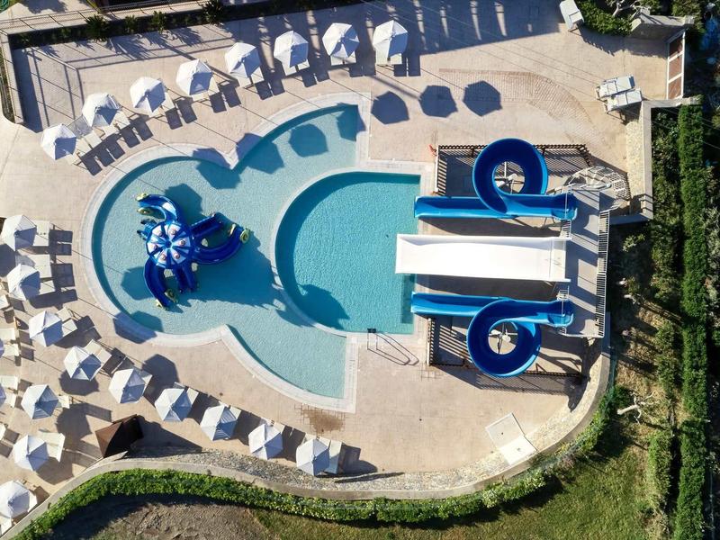 Vista aerea di una piscina a forma di rene con scivoli d'acqua e circondata da lettini e ombrelloni.