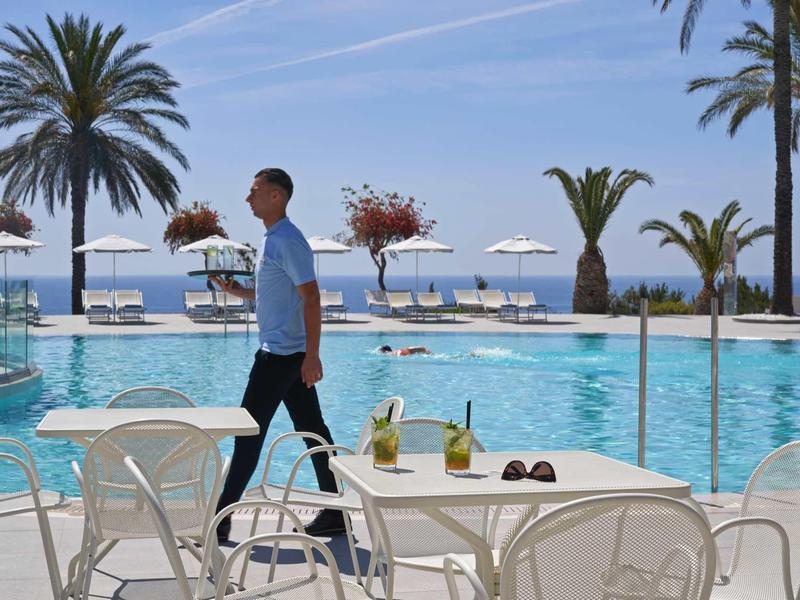 Uomo cammina vicino a tavoli a bordo piscina con bevande e occhiali da sole sotto palme vicino al mare.