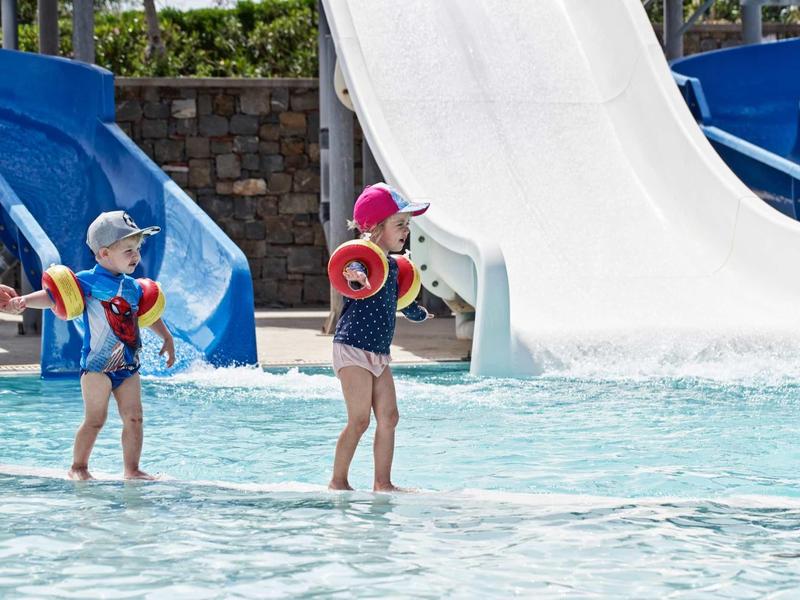 Due bambini che giocano in una piscina bassa con scivoli d'acqua sullo sfondo.