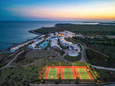 Kustresort met verlichte tennisbanen, zwembaden en gebouwen bij zonsondergang.