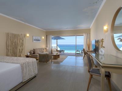 Ruime hotelkamer met zithoek en uitzicht op de oceaan door grote schuifdeuren.