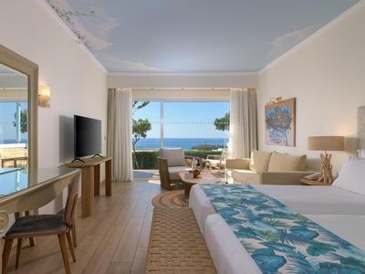 Lichte hotelkamer met groot bed, tv, balkon en zeezicht onder blauwe lucht.