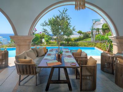Covered terrace dining area overlooking pool and sea under a clear blue sky.