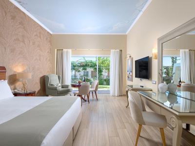 Lichte hotelkamer met tweepersoonsbed, fauteuil, bureau en uitzicht op terras en tuin.