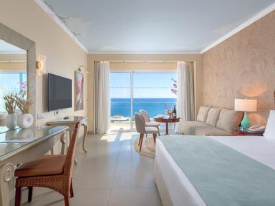 Bright hotel room with sea view, double bed, sofa, table, and TV.