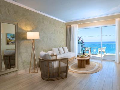 Modern woonkamer met witte bank, rotan stoel en zeezicht door grote ramen.