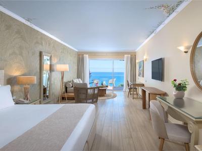 Lichte hotelkamer met tweepersoonsbed, bureau, fauteuils en zeezicht door grote ramen.