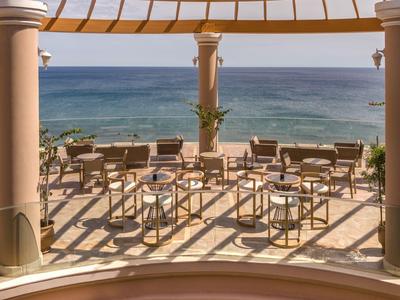 Terrace with tables and chairs under a pergola overlooking the sea on a clear day.