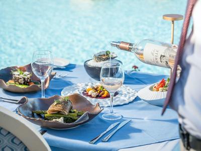 Gedekte tafel met salades en wijn bij een zwembad in de zon.