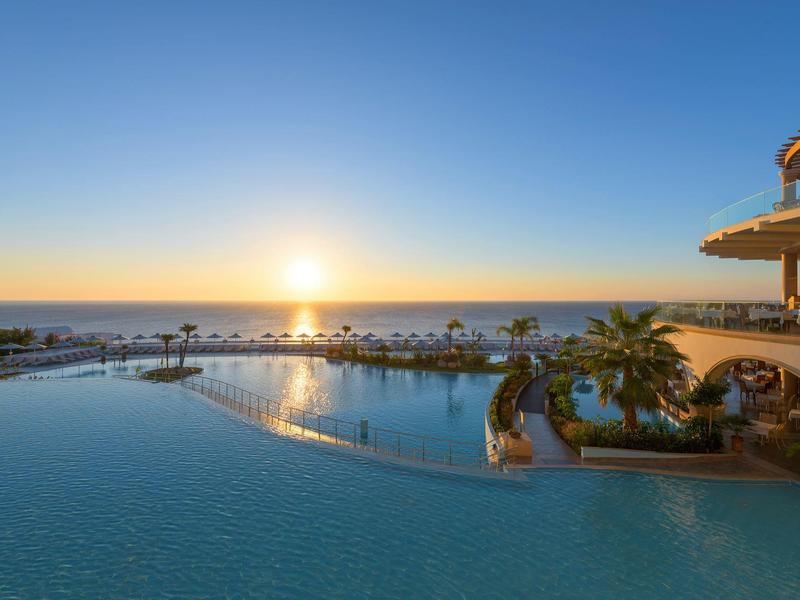 Infinity zwembad met uitzicht op de oceaan bij zonsondergang met palmbomen en resortgebouwen.