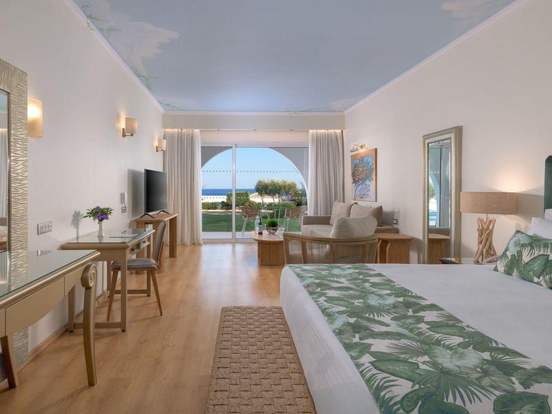 Ruime hotelkamer met groot bed, houten vloer, bureau, zithoek en balkon met zeezicht.