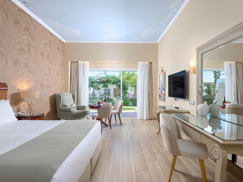 Lichte hotelkamer met tweepersoonsbed, fauteuil, bureau en uitzicht op terras en tuin.