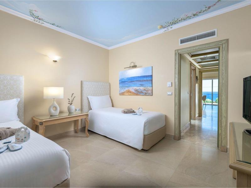 Lichte hotelkamer met twee eenpersoonsbedden, elegante inrichting en uitzicht door de deur op terras en zee.