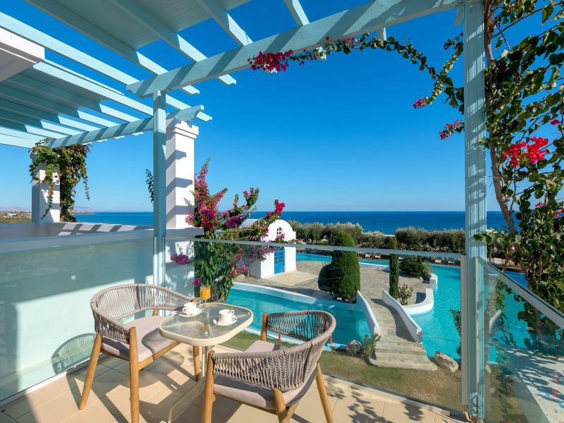 Balkon met stoelen en tafel met uitzicht op zwembad en zee onder een heldere lucht.