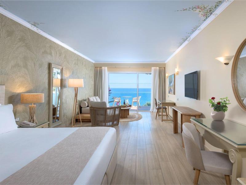 Lichte hotelkamer met tweepersoonsbed, bureau, fauteuils en zeezicht door grote ramen.