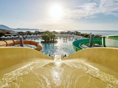 Vista da uno scivolo d'acqua su una piscina con lettini e montagne illuminate dal sole.