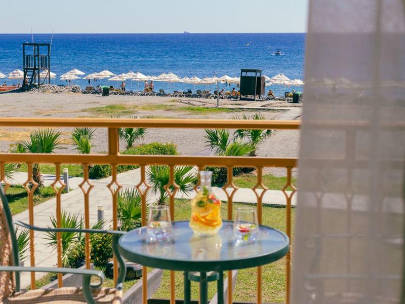 Balkon mit Tisch, zwei Stühlen, Getränk, Blick auf Strand und blaues Meer bei klarem Himmel.