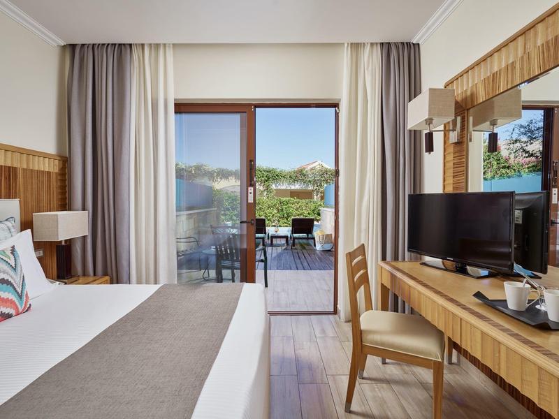 Hell eingerichtetes Hotelzimmer mit Doppelbett, Balkon und Fernseher auf Holzkommode.