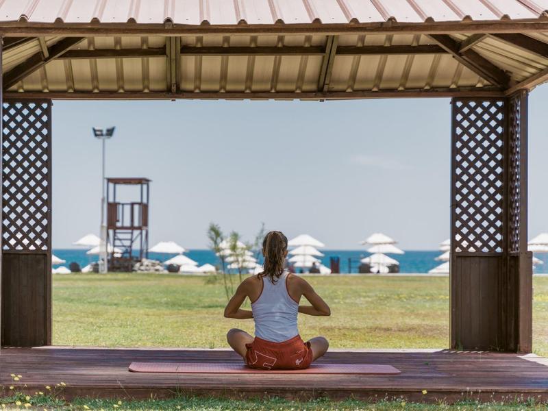 Frau meditiert im Pavillon am Strand, im Hintergrund grüne Wiese, Sonnenschirme und Meer.