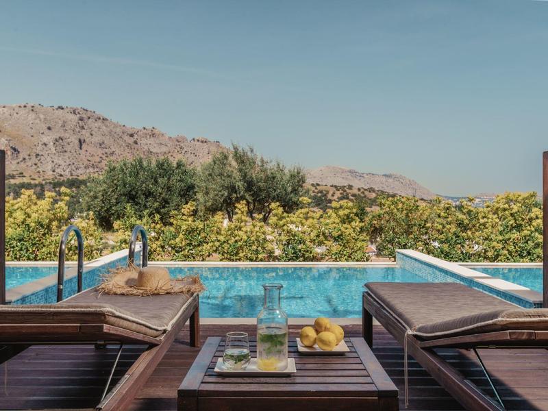 Blick von zwei Holzliegen auf einen Pool mit Obstkorb, umgeben von grünen Bäumen und Bergen im Hintergrund.