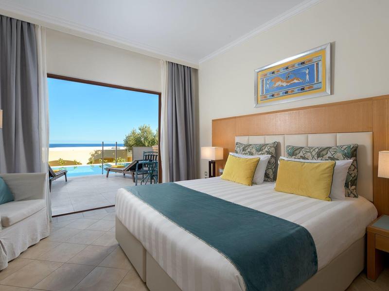 Helles Schlafzimmer mit großem Bett, hellen Vorhängen und Meerblick durch offene Terrassentür.