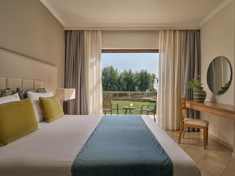 Modernes Schlafzimmer mit großem Bett, grünen Kissen, hellen Vorhängen und Blick auf Garten.