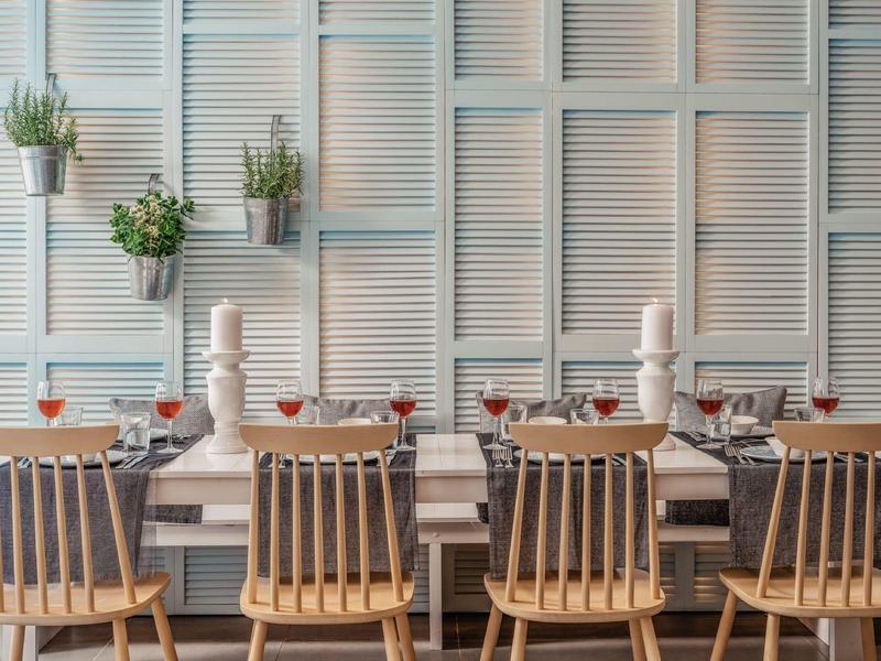 Esstisch mit Holzstühlen, gedeckt mit Gläsern und Kerzen, vor heller Wand mit dekorativen Pflanzen.