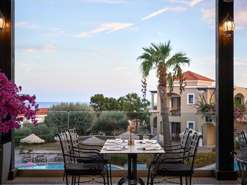 Blick von einer Terrasse mit gedecktem Tisch auf Pool, Palmen und Gebäude bei Sonnenuntergang.