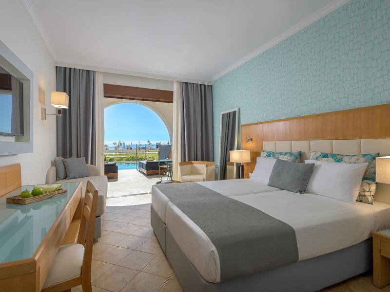 Modernes Hotelzimmer mit großem Bett, Schreibtisch, Fenster zur Terrasse und Meerblick bei Tageslicht.