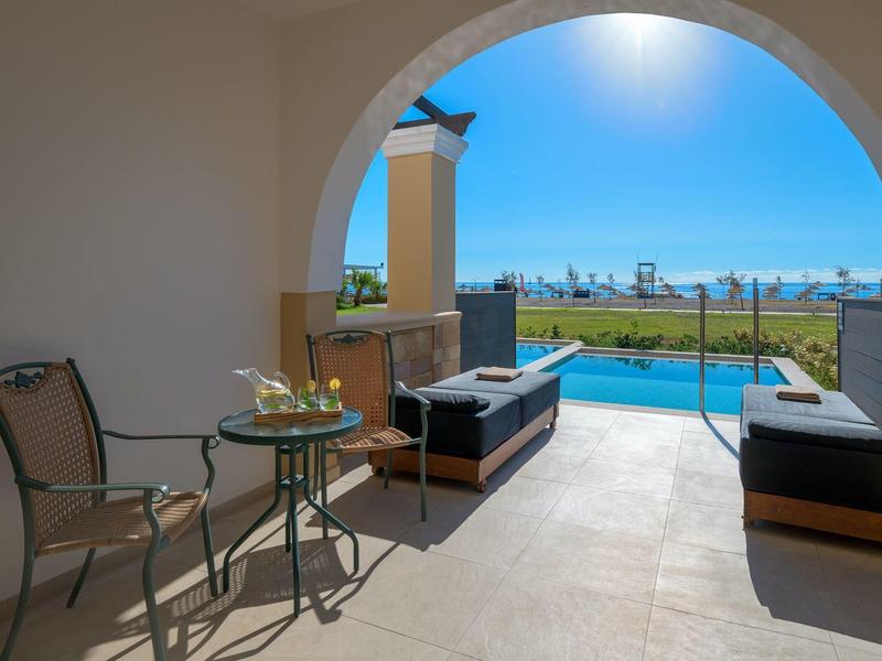 Terrasse mit Sitzgelegenheit, Blick auf Pool und Meer unter blauem Himmel.