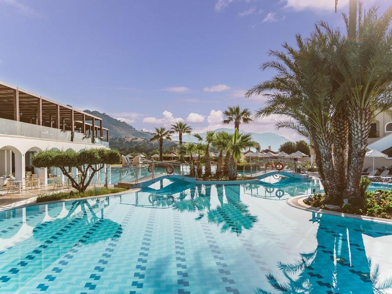 Hotelpool mit Palmen und Bergen im Hintergrund bei sonnigem Himmel.
