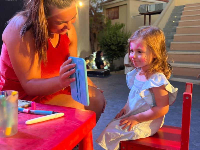 Ein Mädchen sitzt auf einem roten Stuhl, eine Frau zeigt ihm etwas mit einem Tuch an einem roten Tisch.
