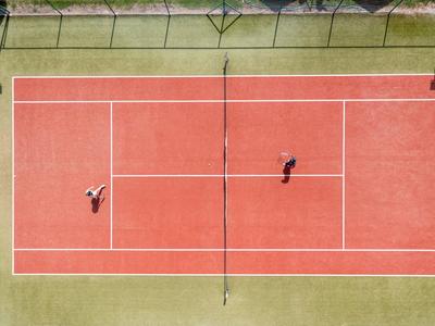 Vista aerea di un campo da tennis rosso con due giocatori e area verde circostante.