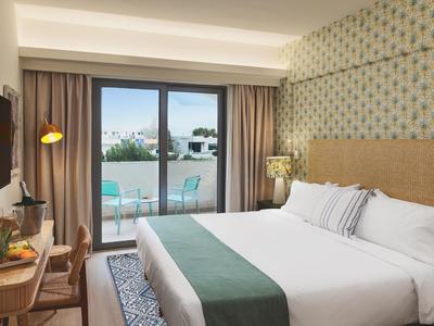 Helles Hotelzimmer mit Doppelbett, Balkon mit Sitzgelegenheit und Stadtblick.