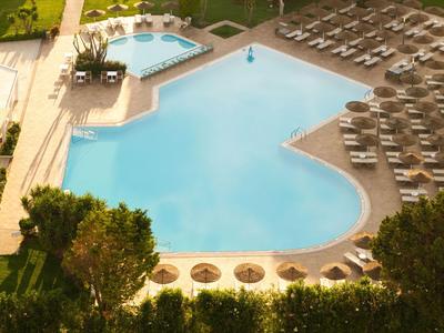 Hotelpool mit Liegestühlen und Bäumen im Sonnenschein.