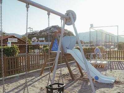 Spielplatz mit Schaukel und Rutsche neben einem Zaun in einem Wohngebiet bei klarem Himmel.