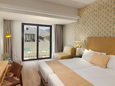 Helles Hotelzimmer mit großem Bett, Sesseln und Blick auf einen Pool und Berge.